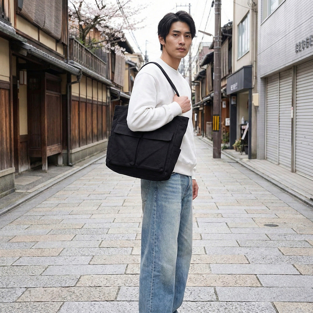 Man in a white sweatshirt and light jeans carries the black tote on one shoulder while standing on a paved street. 2