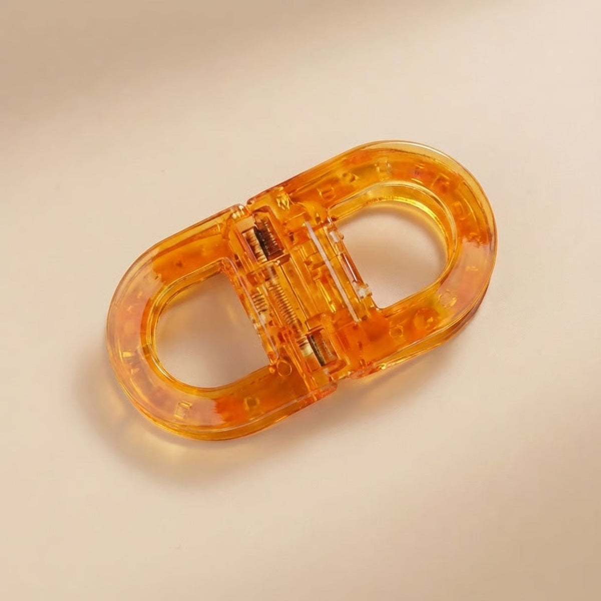 Translucent amber oval hair clip with visible spring mechanism, shown flat from above on a soft beige background. 1