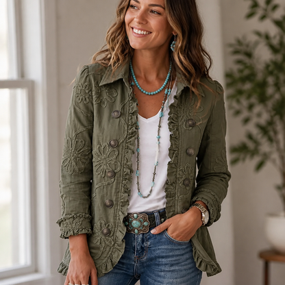 Smiling woman in an olive green embroidered jacket with ruffle edges, worn open over a white tee and jeans beside a window. 1