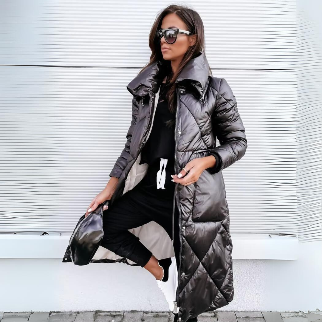Woman in sunglasses wears the black quilted long coat open over black loungewear, holding a small black bag against a pale ribbed wall. 1