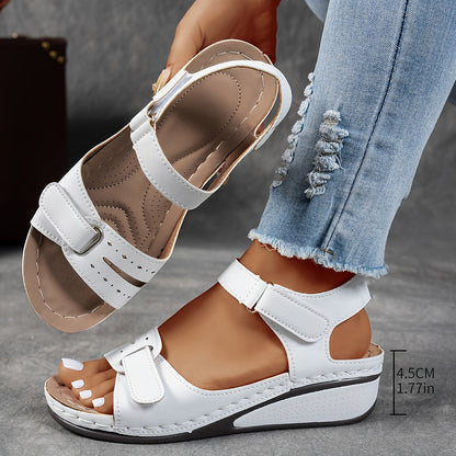 White wedge sandals with hook-and-loop straps, worn by a model while the other sandal is held up to show its cushioned footbed. 1