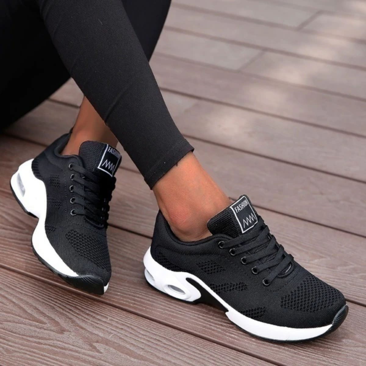 Black mesh trainers with white soles worn by a model in black leggings, feet crossed on wooden decking. 8
