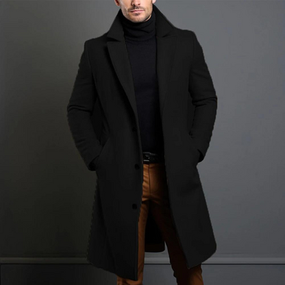 Model wearing the black single-breasted longline coat with notch lapels over a black roll-neck jumper and tan trousers, hands in pockets. 1