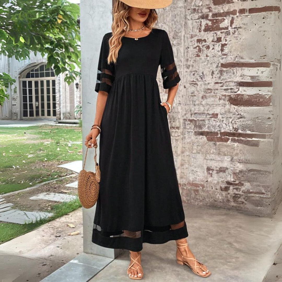 Model wearing the black maxi dress outdoors with a straw hat, woven round bag and strappy sandals, one hand in a pocket. 1
