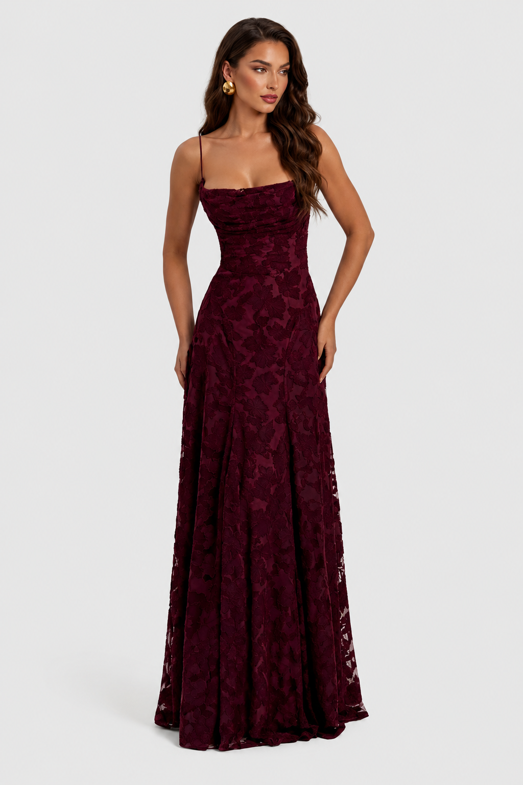 Model in burgundy floral lace gown with draped cowl neckline, front view, hands resting at her hips, wearing gold earrings. 9