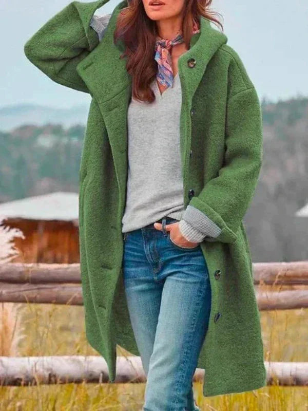 Woman wearing the long hooded coat in green over a grey jumper and jeans, with a scarf, standing by a wooden fence. 5