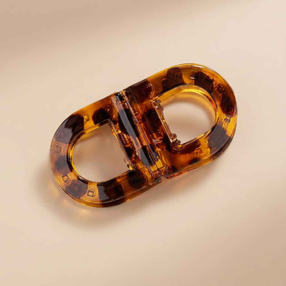 Flat clip in glossy tortoiseshell with dark brown and amber patches, photographed from above on a beige surface. 4