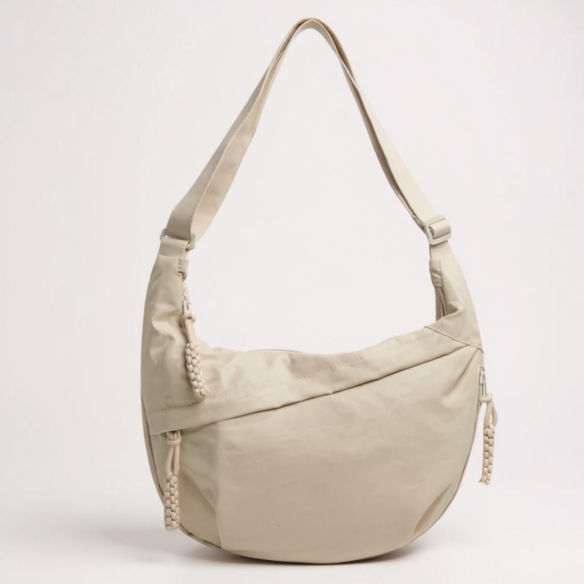 Cream crescent shoulder bag from the front, with braided zip pulls and a matching adjustable strap, on a plain pale background. 6
