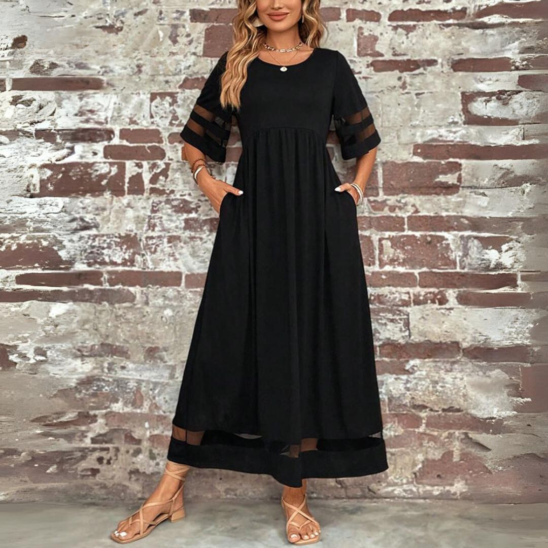 Front view of the black maxi dress with sheer striped sleeves and hem, worn against a brick wall, hands in pockets. 2