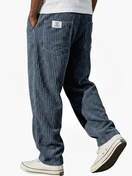 Back view of blue-grey wide-wale corduroy trousers worn with a white tee and white trainers, showing the back pocket and woven patch. 2