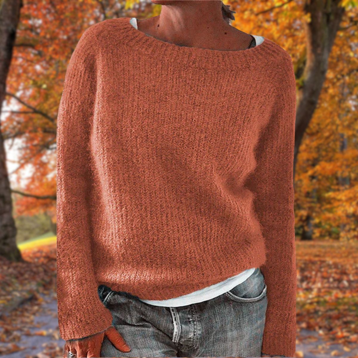 Rust orange knit sweater with a wide neckline, worn over a white top and grey jeans among autumn leaves 6