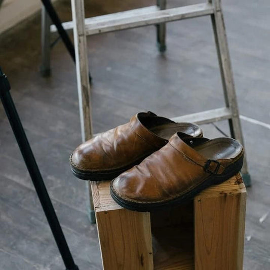 Pair of worn tan leather backless clogs resting on a wooden block beside a stepladder 1