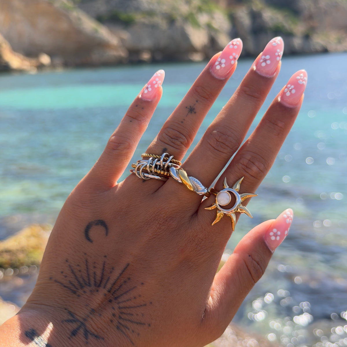 Hand held up against a turquoise sea wearing the stacked ring on the middle finger beside a sun-shaped ring. 3