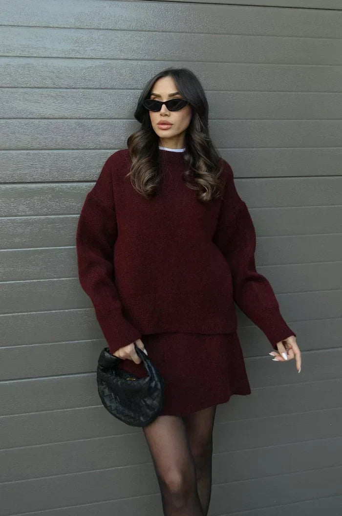 Model in burgundy knit sweater and skirt, holding a black woven clutch, leaning against a grey wall 1