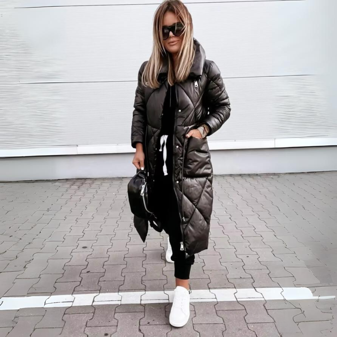 Model in the black quilted long coat with a high collar and hands in the side pockets, standing on paving. 5