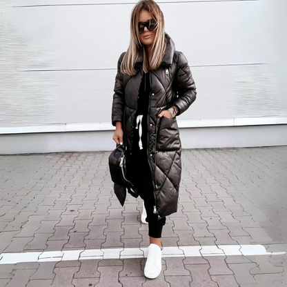 Model in the black quilted long coat with a high collar and hands in the side pockets, standing on paving. 5