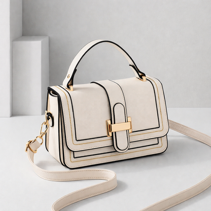 Cream bag with top handle, gold clasp and contrast stitching, shown at an angle with its long strap curled on a pale studio floor. 1