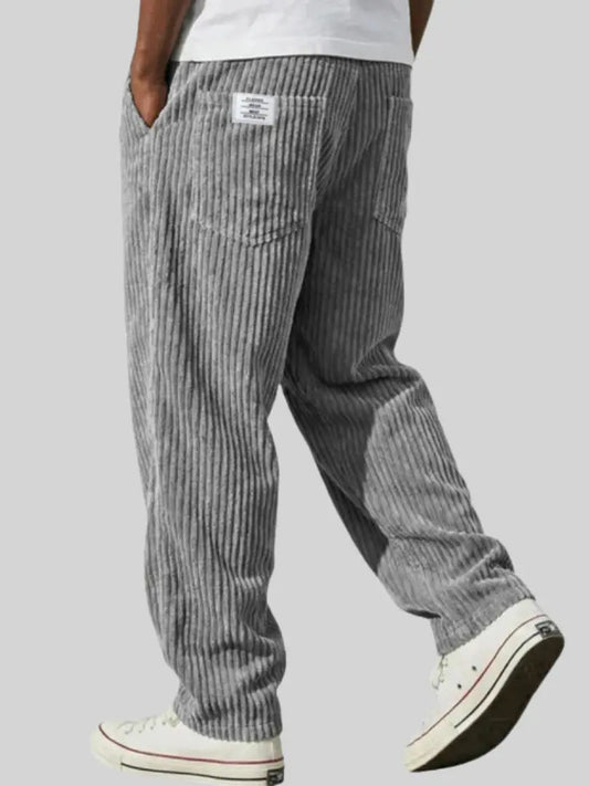 Back view of grey wide-wale corduroy trousers worn by a model with a white tee and white trainers, one hand in pocket. 1