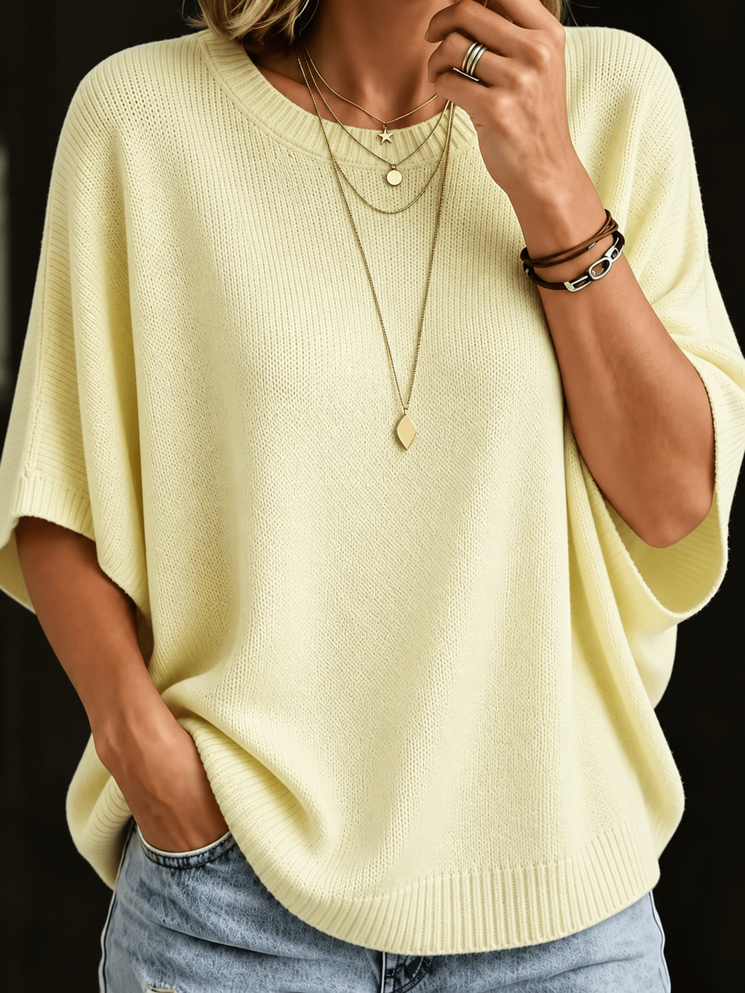 Close-up of the pale yellow knit batwing top with ribbed round neck, worn with layered gold necklaces and jeans. 4