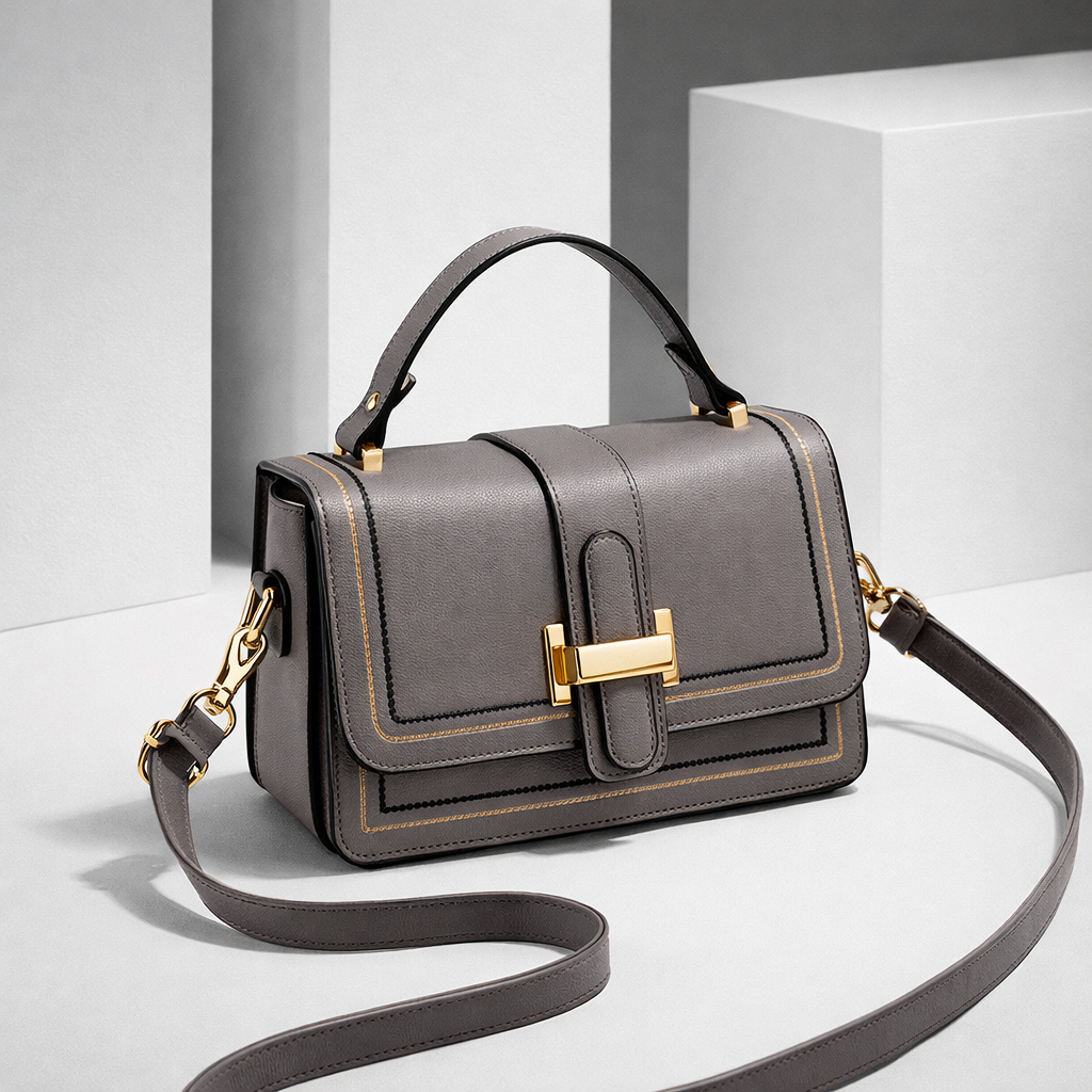 Grey version of the bag at an angle on a pale studio set, with gold clasp, gold stitching and strap curled in front. 3