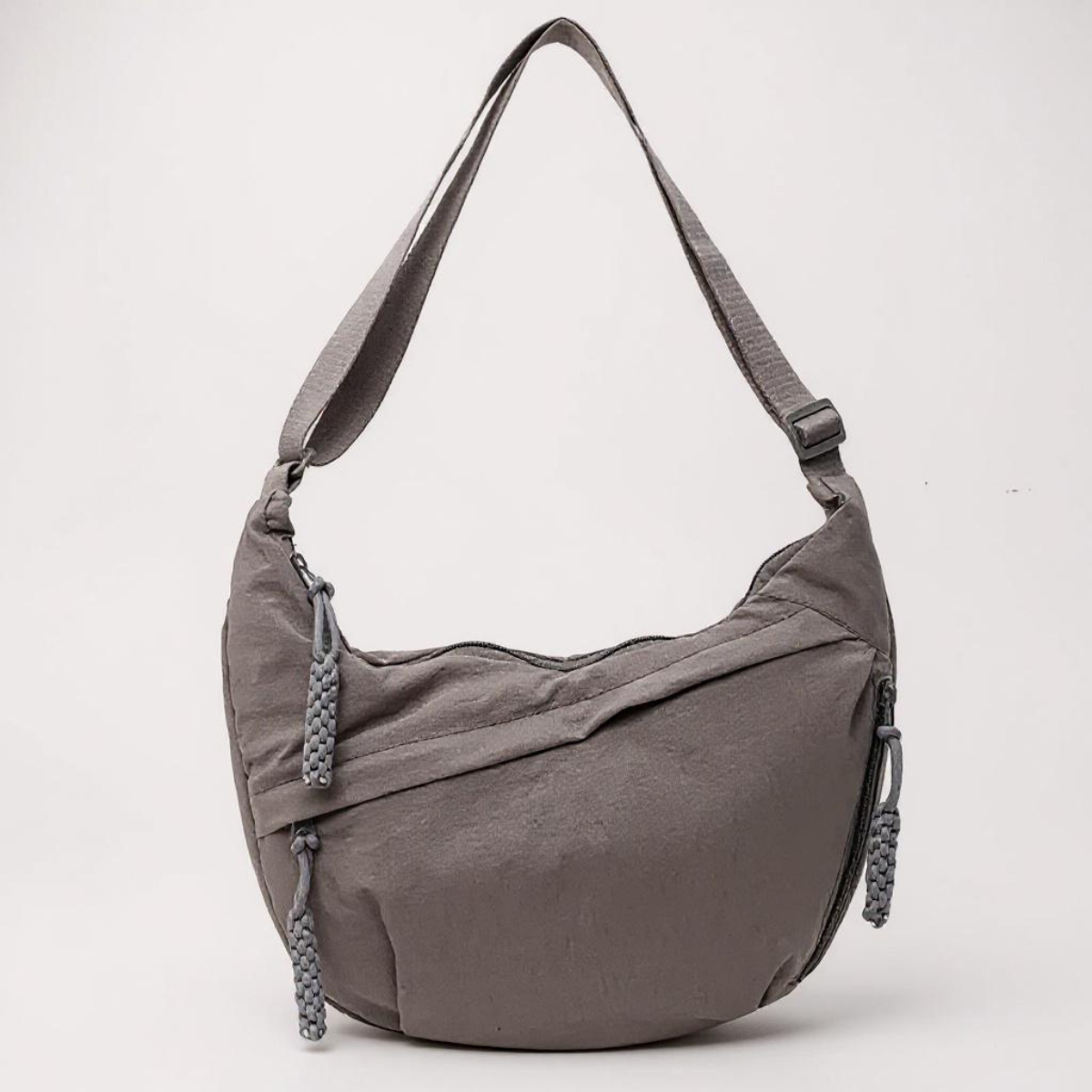 Crescent-shaped nylon shoulder bag in taupe grey against a pale background, with adjustable strap and braided zip pull tassels. 7