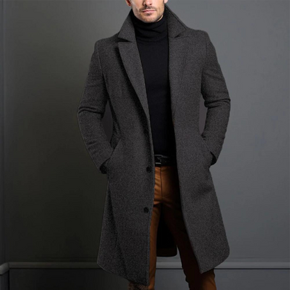 Model wearing the dark grey, textured version of the long tailored coat with a black roll-neck, hands in the pockets. 3