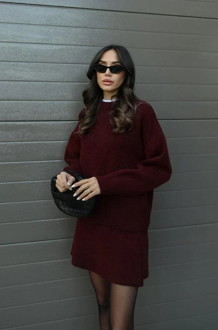 Model in the burgundy set, three-quarter pose, hands clasped on a black woven handbag 3