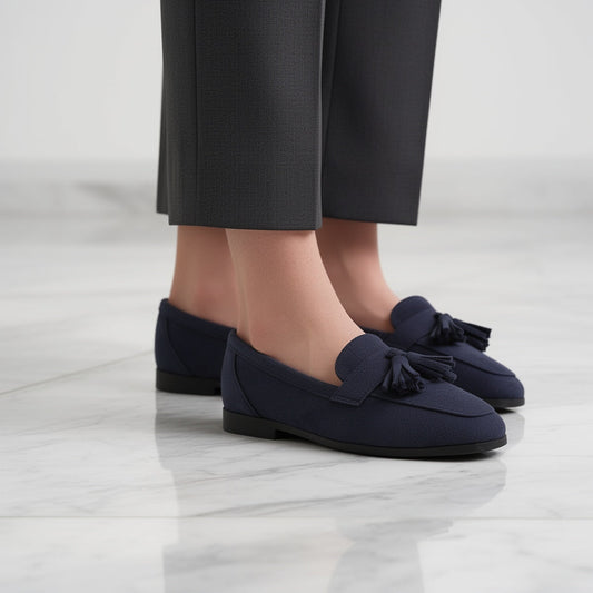 Navy tasselled loafers worn with grey trousers, side view on a white marble floor 1