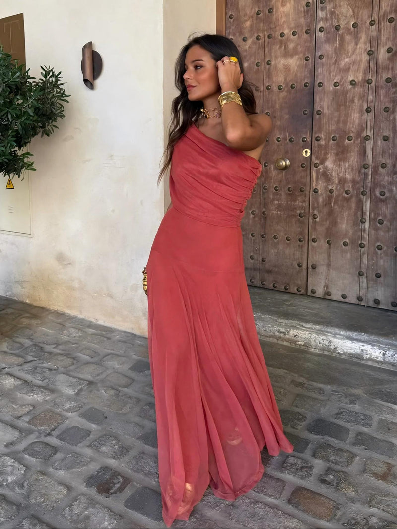 Model in terracotta one-shoulder mesh dress, hand in hair, standing by a studded wooden door 4