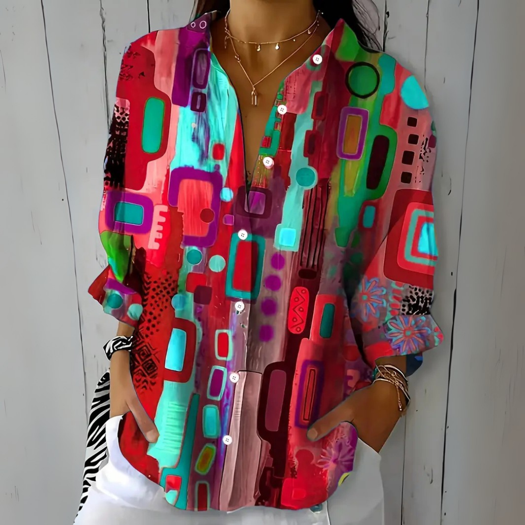 Model wearing the blouse in a red and pink colourway with turquoise and purple abstract shapes, one hand in a trouser pocket. 4