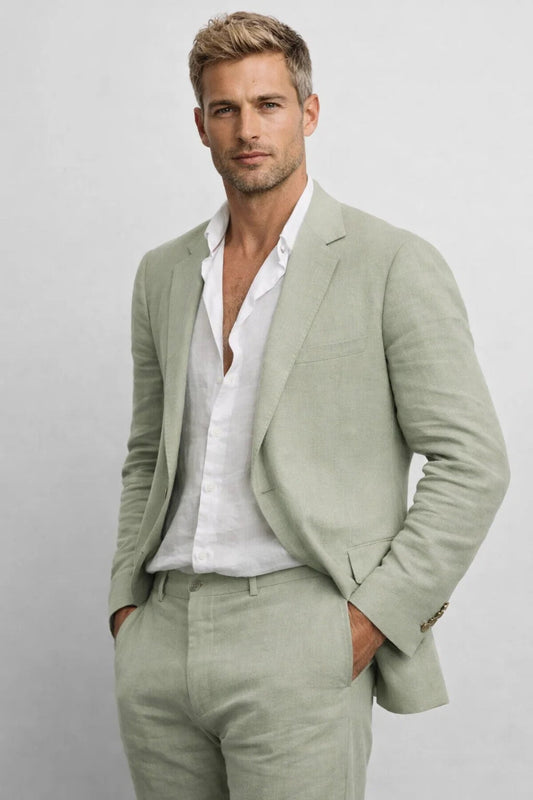 Model in a sage green suit with an open-necked white shirt, hands in pockets, shown from the thighs up. 1