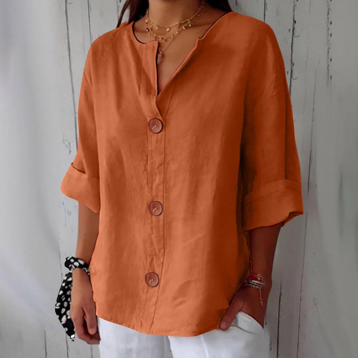 Model wearing the loose blouse in burnt orange, with a V-neckline, three round buttons and folded sleeve cuffs, paired with white trousers. 7