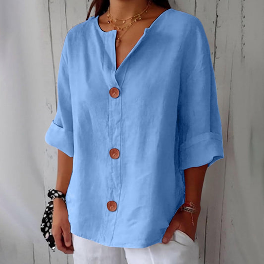 Model wearing the loose blouse in light blue with three round wooden-look buttons, rolled sleeves and a notched V-neck, with white trousers. 1