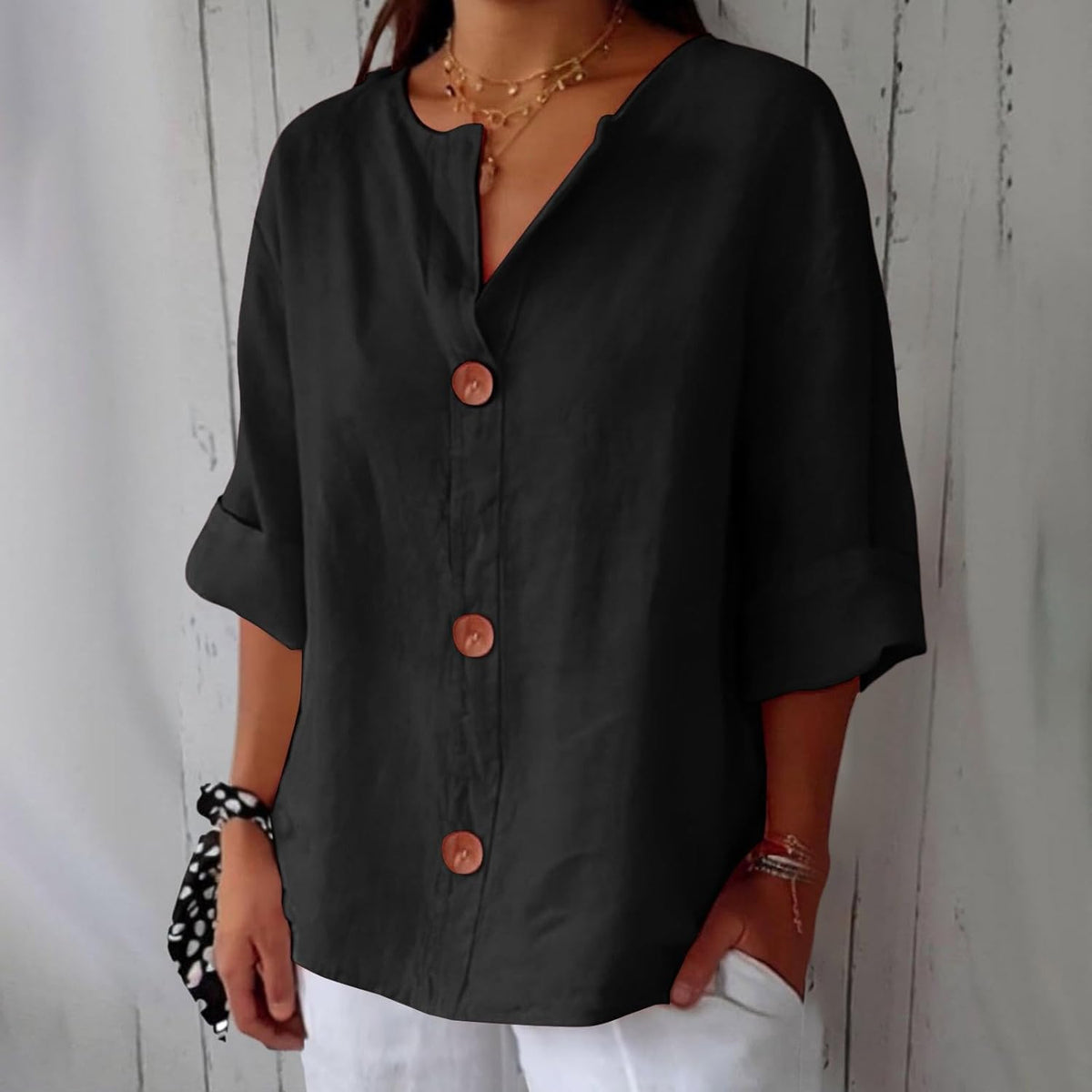 Model wearing the loose button-front blouse in black, with brown buttons, rolled sleeves and layered gold necklaces against a whitewashed wooden wall. 11