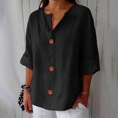 Model wearing the loose button-front blouse in black, with brown buttons, rolled sleeves and layered gold necklaces against a whitewashed wooden wall. 11