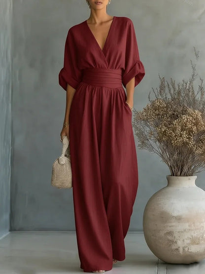 Burgundy wide-leg jumpsuit in full-length studio view, with wrap V-neck, wide waistband and rolled sleeves, beside a dried-flower vase. 13