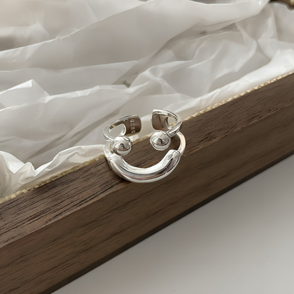 Silver ring resting on a wooden tray lined with white tissue paper, its smiling face and small stamp inside the band visible. 6