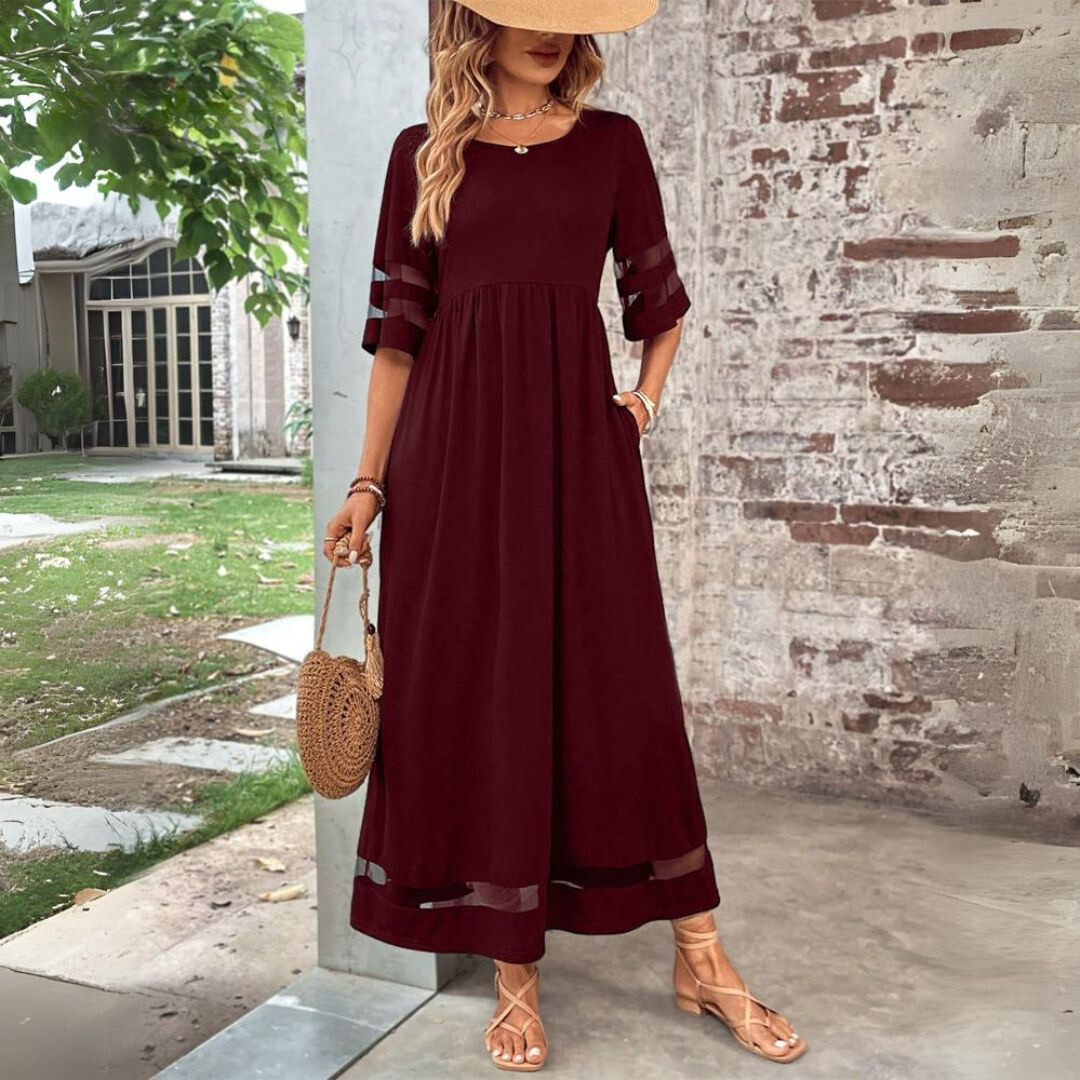 Burgundy version of the maxi dress worn with a straw hat and woven round bag, standing outdoors beside a brick wall. 5