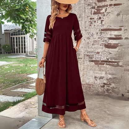 Burgundy version of the maxi dress worn with a straw hat and woven round bag, standing outdoors beside a brick wall. 5