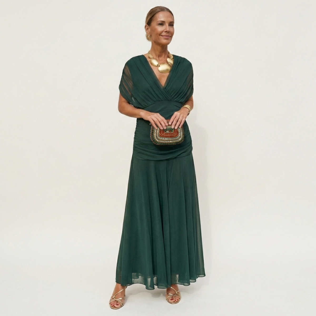 Model in a full-length dark green chiffon dress with a ruched V-neck bodice and sheer cap sleeves, holding a beaded clutch. 1