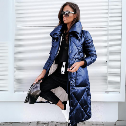The long quilted coat in glossy navy blue, worn open over a black outfit and white trainers by a model in sunglasses. 3