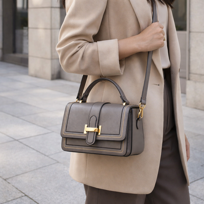 Model in a beige coat wearing the grey bag across the body, with its top handle and gold clasp on show. 4