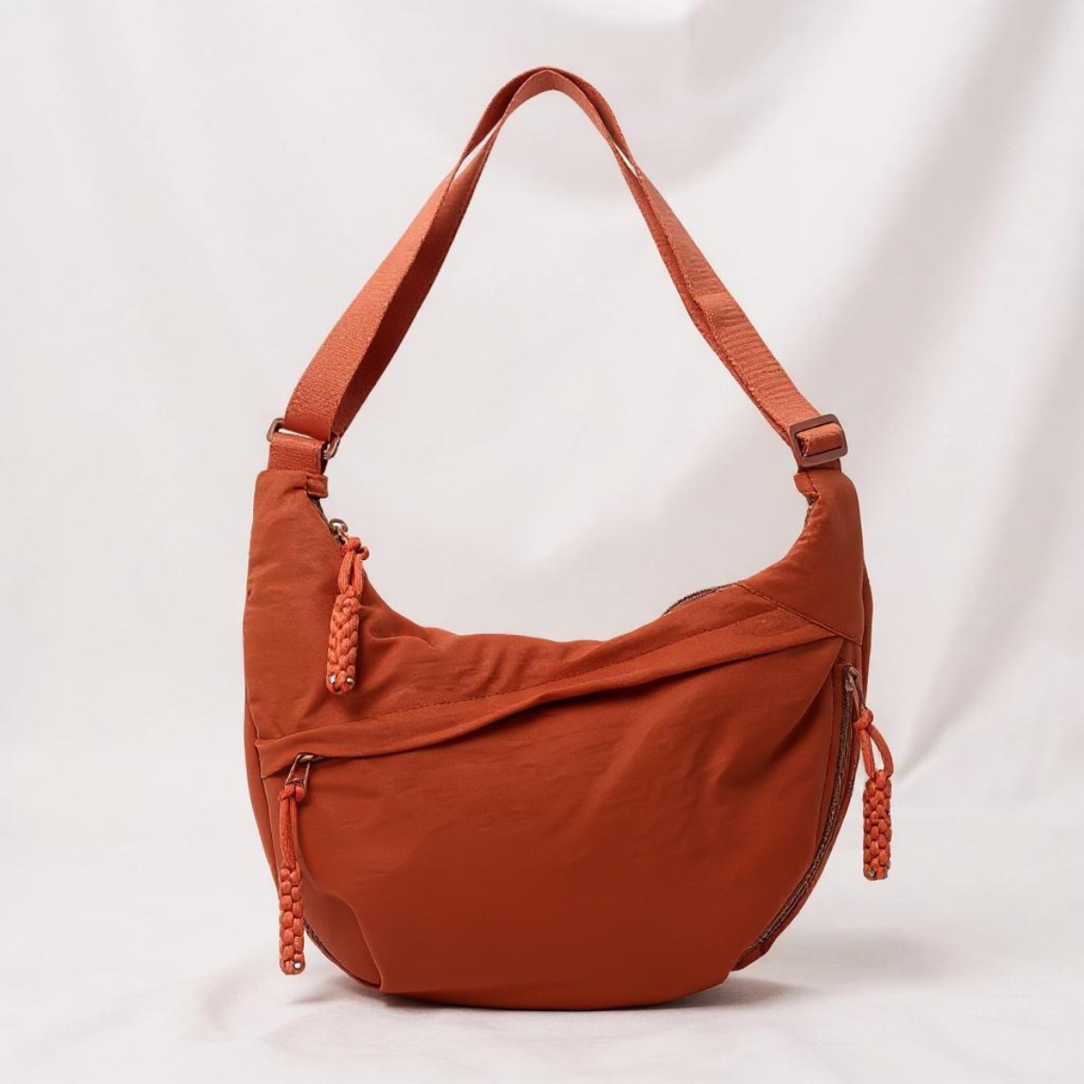 Crescent-shaped nylon shoulder bag in rust orange on a pale draped backdrop, with adjustable strap and braided zip pulls. 2