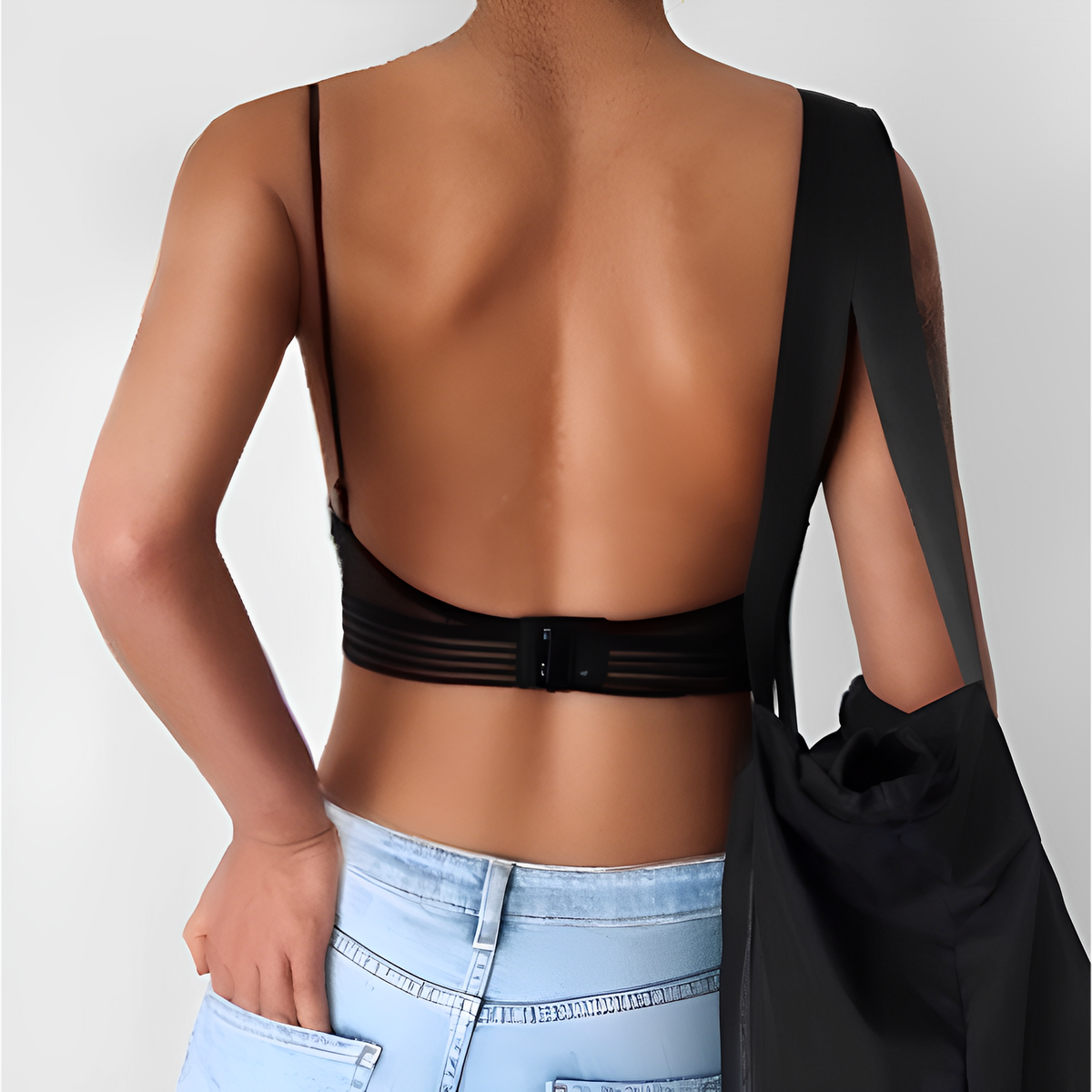 Back view of a model wearing a black bralette with a low scooped back and striped elastic band, carrying a black tote. 2