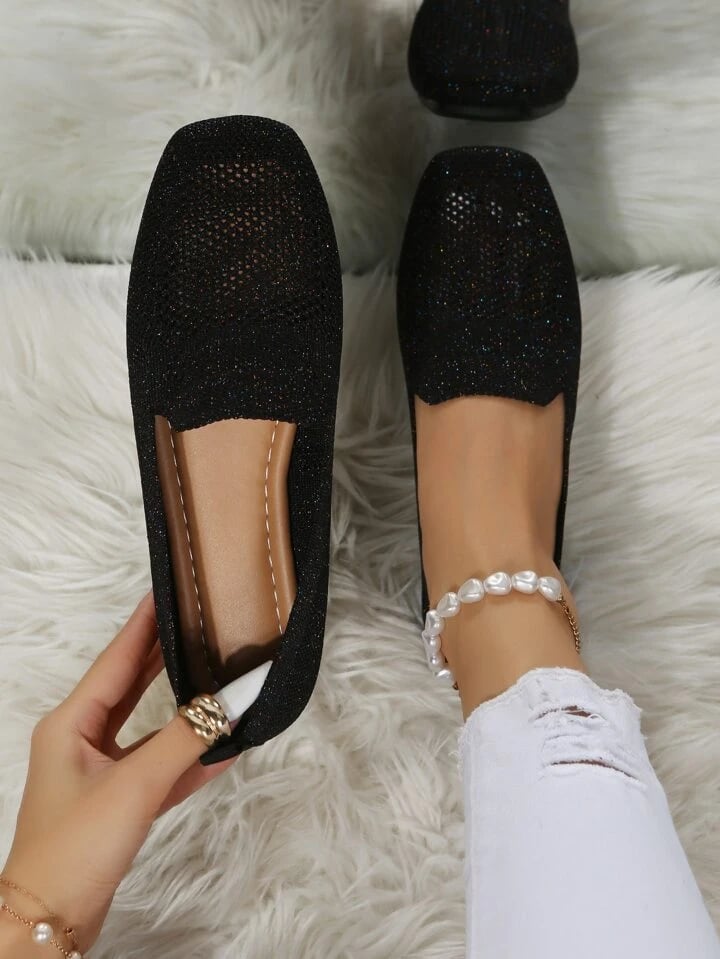Black glittery mesh slip-ons seen from above on a fluffy white rug, one held to show the soft footbed. 1