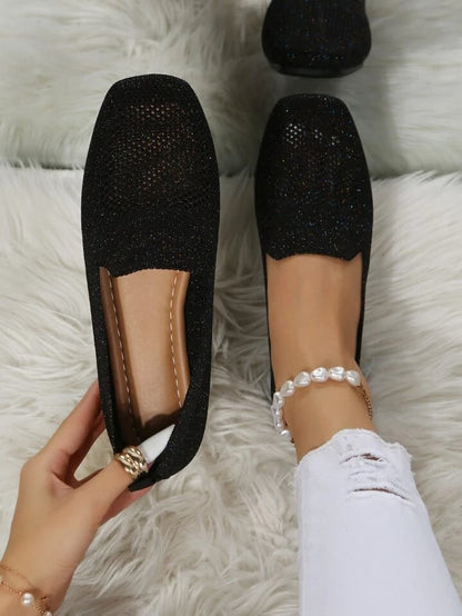 Black glittery mesh slip-ons seen from above on a fluffy white rug, one held to show the soft footbed. 1