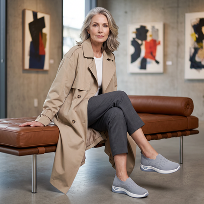Grey slip-on trainers with white soles worn by a woman in a beige trench coat, sitting on a leather bench. 8