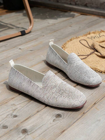 Pair of light grey knit slip-ons with white heel pull tabs standing on wooden boards beside a straw mat. 17