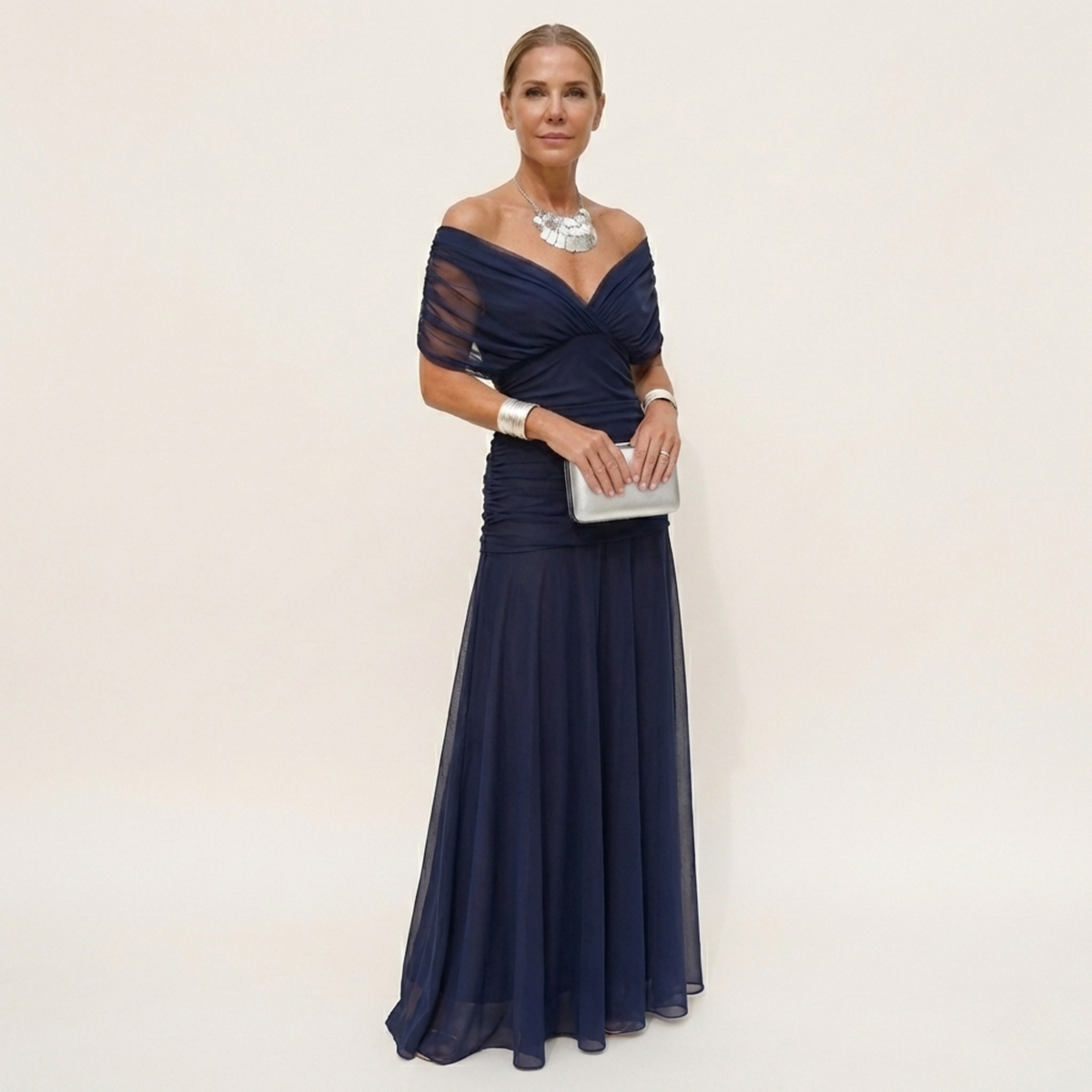 Full-length view of a navy chiffon version with an off-the-shoulder ruched bodice, worn with a silver clutch and necklace. 3