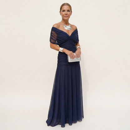 Full-length view of a navy chiffon version with an off-the-shoulder ruched bodice, worn with a silver clutch and necklace. 3
